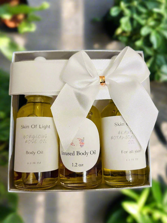 A clear gift box containing three travel size bottles of body oil with a white ribbon bow on top. The bottles are labeled Skin of Light, Gorgeous Rose, and Infused Body Oil.